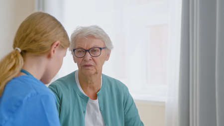 Senior lady patient with glasses talks to experienced young caretaker sitting near window in light room at long term care homeの写真素材