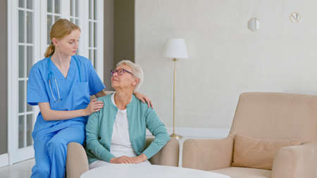 Smiling blonde nurse in uniform takes care of senior woman sitting in comfortable armchair in light room at assisted livingの写真素材