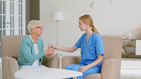 Young blonde doctor in uniform gives glass of water to senior patient woman at small table in light room at long term care homeの写真素材