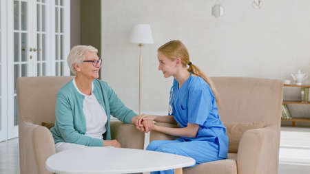 Positive doctor joins hands cheering up mature gray haired lady sitting in armchairs in stylish office at long term care homeの写真素材