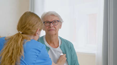 Senior woman patient undergoes examining with stethoscope and doctor listens to heartbeat near window in at long term care homeの写真素材