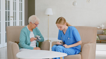 Young girl in blue uniform puts digital blood pressure monitor on senior woman hand in light office at homeの写真素材