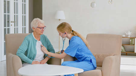 Young nurse measuring heart rate with stethoscope in stylish office at long term care homeの写真素材