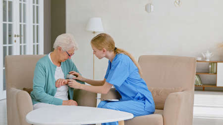 Young nurse in blue uniform puts digital blood pressure monitor on senior woman hand in light office at long term care homeの写真素材