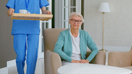 Lady medical worker in blue uniform carries drink and fruits to old woman patient sitting in armchair at long term care homeの写真素材