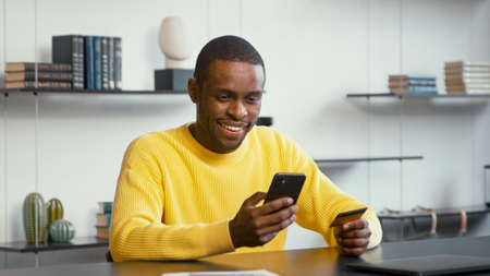 Smiling man in yellow shirt looks at bank card in hand and enters digits to smartphone sitting at...