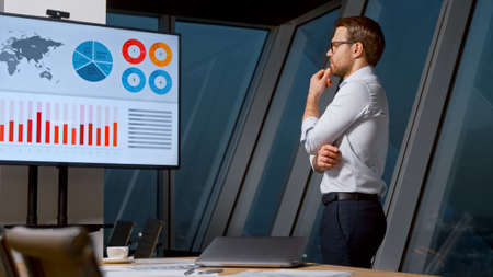 Thinking young man looking at screen with analytics in meeting room in the eveningの写真素材