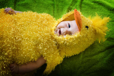 Funny infant in yellow chicken costume lies on the green plain and looks at the camera with happy smileの写真素材