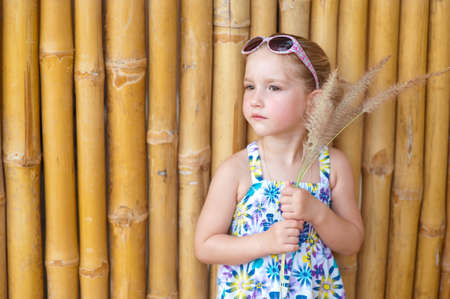 Sad blond toddler gerl with grass near bamboo wall in sunglassesの写真素材