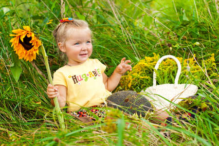 Adorable blond girl sitting in grass with sunflowers and smilingの写真素材