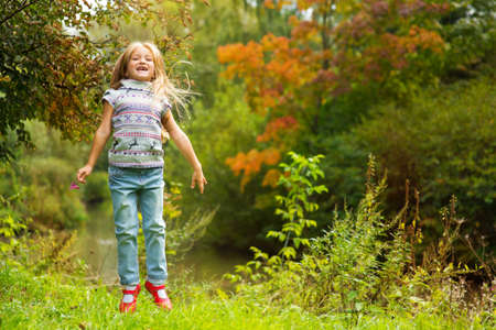 Funny toddler european girl with long blond hair jumping on the river shore in colorful autumn forestの写真素材
