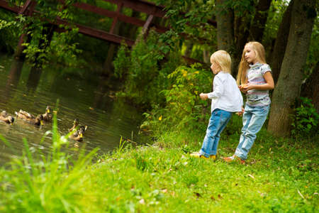 Two cute european sisters standing on forest river shore and feeding ducks with breadの写真素材