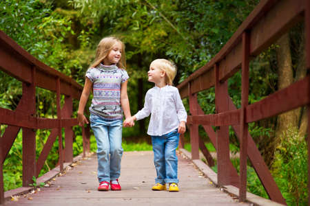 Two cute european blond sisters on scenic bridge together having fun with colorful summer forest on backgroundの写真素材