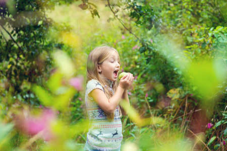 Cute european blond girl ready to eat green apple in colorful parkの写真素材