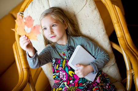 Beautiful european toddler girl sitting on armchair with yellow maple leaf with hole like book in it   Girl holding a book の写真素材