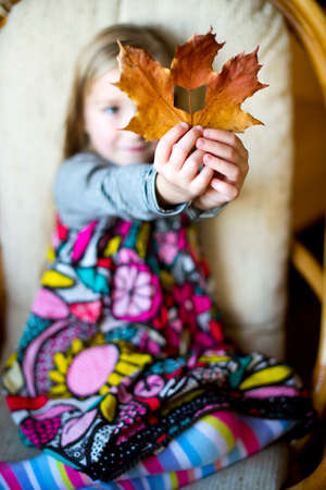 Beautiful european toddler girl sitting on armchair with yellow maple leaf with hole like book in itの写真素材