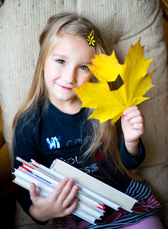 Beautiful european toddler girl sitting on armchair with yellow maple leaf with hole like book in it holding booksの写真素材