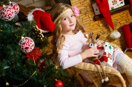 Beautiful toddler european girl with long blond curly hair sitting near fireplace with present box and celebrates christmas under xmas treeの写真素材
