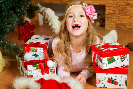 Happy european girl with long blond curly hair and flower on it lies under Christmas tree with her presents and screams gailyの写真素材