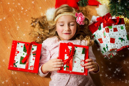 Happy european girl with long blond curly hair and flower on it lies under Christmas tree with her presents and closed eyes, making her wishの写真素材