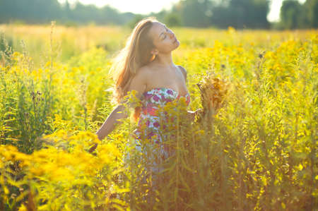 Happy european young woman in summer blooming field holding yellow flowers and walking under evening sunの写真素材