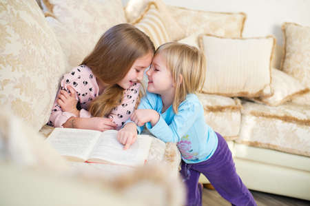 Two cute european sister girls reading a book for children and having fun in room of their homeの写真素材