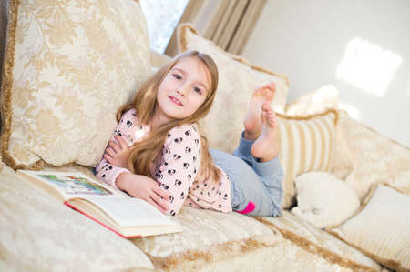 One cute toddler european girl with long blond hair reading book on sofa in the room at homeの写真素材