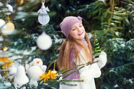 Happy european toddler girl outdoors near xmas tree, decorated with holiday toys making her wish with smile and closed eyesの写真素材