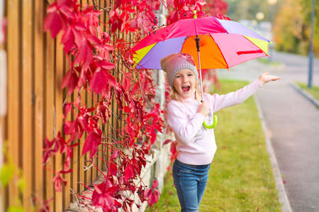 Funny toddler girl with colorful umbrella stay in red leaf autumn garden and smileの写真素材