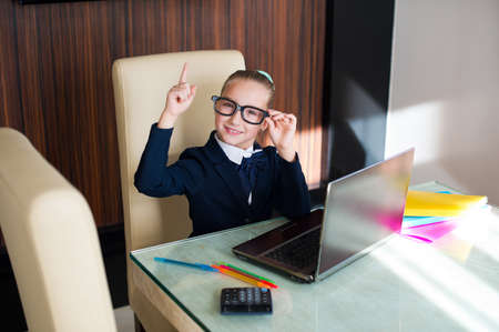Beautiful toddler smart school girl in glasses sitting at table with laptop doing her home workの写真素材
