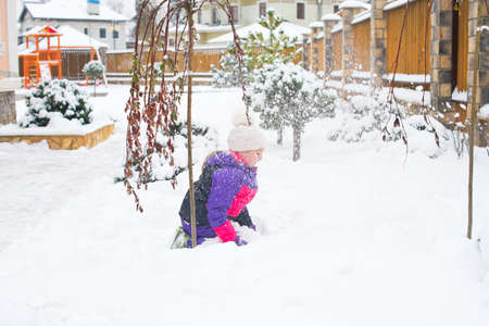 Happy little girl in colorful suit and white hat play with snow in back yard of village country house in cold winter weatherの写真素材