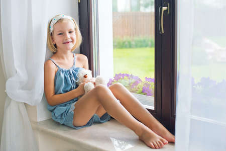 Lonely toddler russian girl sitting near window at home playing teddy bears happy and funny. Colorful back yard at backgroundの写真素材