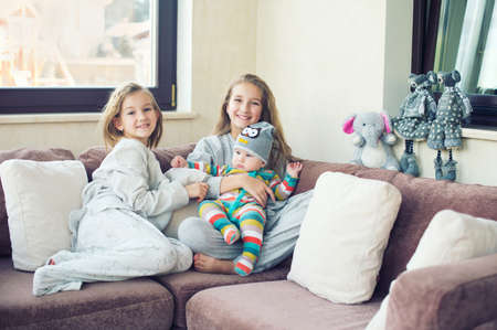 Two adorable european sisters girls holding theur newborn brother having fun in livingroom of theis home. Family with three kids.の写真素材