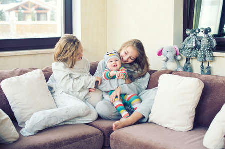 Two adorable european sisters girls holding theur newborn brother having fun in livingroom of theis home. Family with three kids.の写真素材