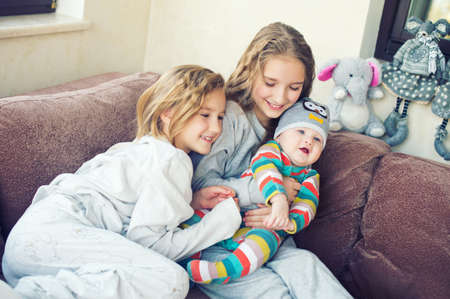 Two adorable european sisters girls holding theur newborn brother having fun in livingroom of theis...