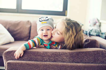 Beautiful toddler girl kissing her little brother at home, laughing and embracing with love and careの写真素材