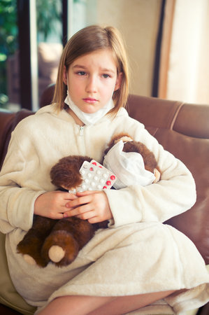Little toddler girl sitting in medical mask with teddy bear feeling sick with flu and fever, holding pills and ready to take a cureの写真素材