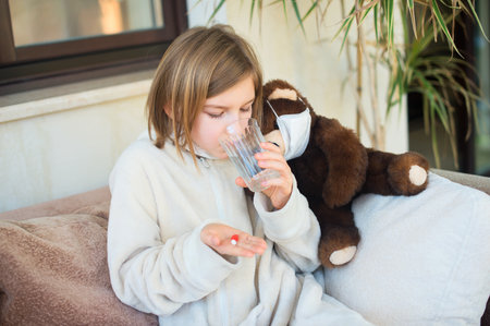 Sad sick toddler girl with fever in medicine mask ready to take cure pills and get well soon from her illnessの写真素材