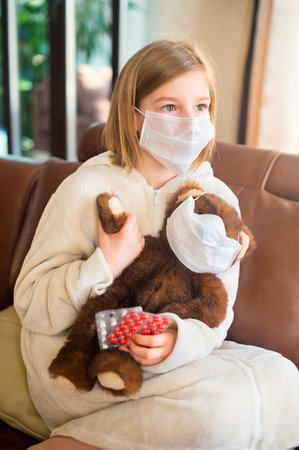 Little toddler girl sitting in medical mask with teddy bear feeling sick with flu and fever, holding pills and ready to take a cureの写真素材