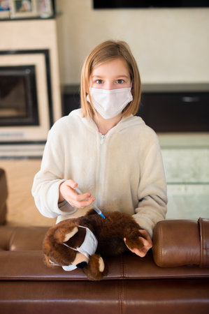 Sick toddler girl in medicine mask making vaccination her teddy bear with syringe. Playing in doctorの写真素材