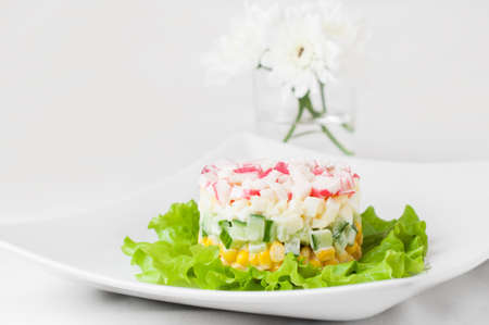 Layered salad of crab sticks, canned corn, cucumber and eggs on lettuce leaves and white flowers on a white tableclothの写真素材