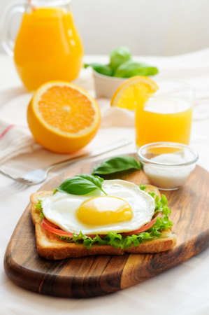 Breakfast of toast with fried egg, tomato and green salad, decorated with basil leaves on a wooden cutting board and jug of orange juiceの写真素材