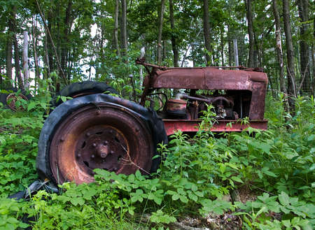 Rusting, broken, old tractor with flat tires sitting in a field, overgrown with green vegetationの写真素材