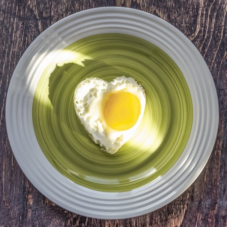 Fried egg in heart shape on a platter on wooden background. Sunlight on the dishの写真素材