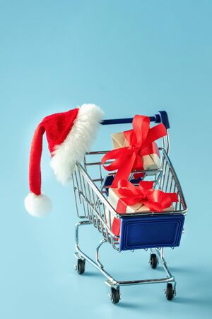 Mini trolley with gift boxes and red Santa Claus cap on blue backgroundの写真素材