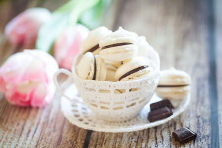 Vanilla macarons with chocolate ganache in a white knit cup and pink tulips on wooden tableの写真素材