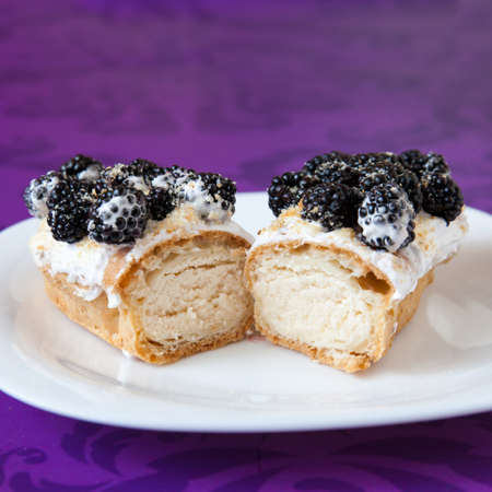 Cut eclair with cream and blackberies on a white plate on violet background