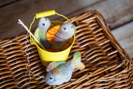 Beautiful colored felted fish with fishing rod in a small yellow pail on wooden basketの写真素材