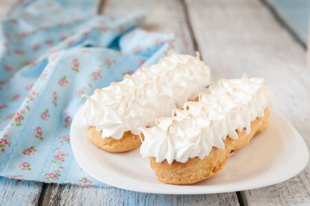 Eclairs with meringue frosting on a white plate with fabric on light wooden background