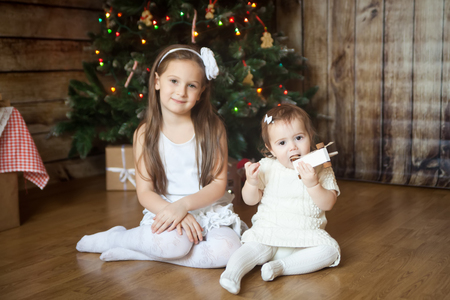 Two cute sisters in white dresses sitting in front of decorated Christmas treeの写真素材